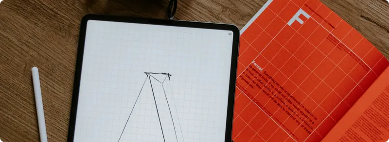 iPad with a wireframe letter sketch and a pencil next to an open orange typography book on a wooden desk