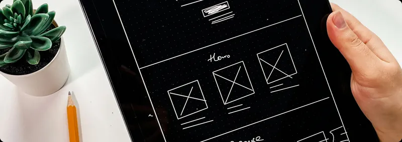 Hand holding a tablet with a hand-drawn website wireframe next to a succulent and a pencil