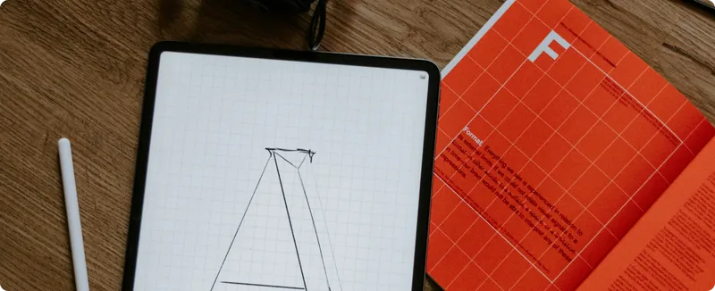 iPad showing a sketched letter A beside an orange type specimen book on a wooden desk